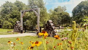 A rustic outdoor setting with wooden barrels stacked under a wooden archway, surrounded by lush greenery and wildflowers.