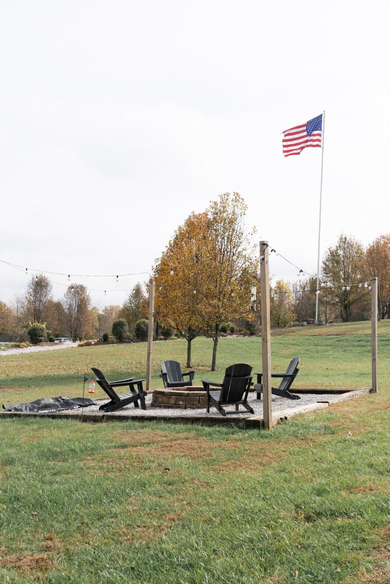 A grassy outdoor area with a flagpole, a fire pit, and several Adirondack chairs.