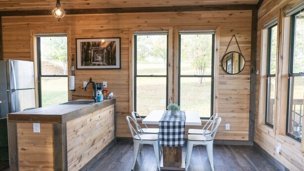 A wooden cabin interior with a kitchen area, dining table, and large windows. - Bourbon Barrel Cottages #3 on Ky. Bourbon Trail (Lawrenceburg)