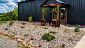 A building with a dark exterior, a wooden porch, and a gravel garden with flowers. - Bourbon Barrel Cottages #3 on Ky. Bourbon Trail (Lawrenceburg)