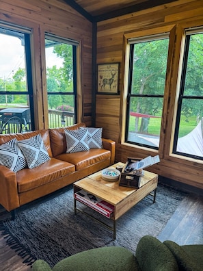 Interior de una cabaña de madera con sofá de cuero, una mesa de centro y ventanas amplias.