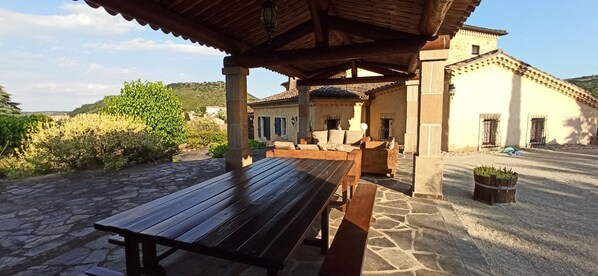 A covered outdoor seating area with stone flooring, a wooden table, and a wicker sofa.