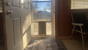 A room with a wooden door, a view through the door, a window with blinds, and a chair. - Rose District Bungalow pet friendly (Broken Arrow)