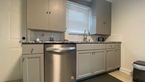 A kitchen with grey cabinets, a stainless steel dishwasher, and a window with blinds. - Rose District Bungalow pet friendly (Broken Arrow)