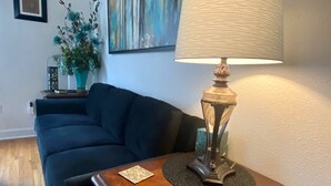 A living room with a dark blue sofa, a wooden side table with a lamp, a painting on the wall, and a potted plant. - Rose District Bungalow pet friendly (Broken Arrow)