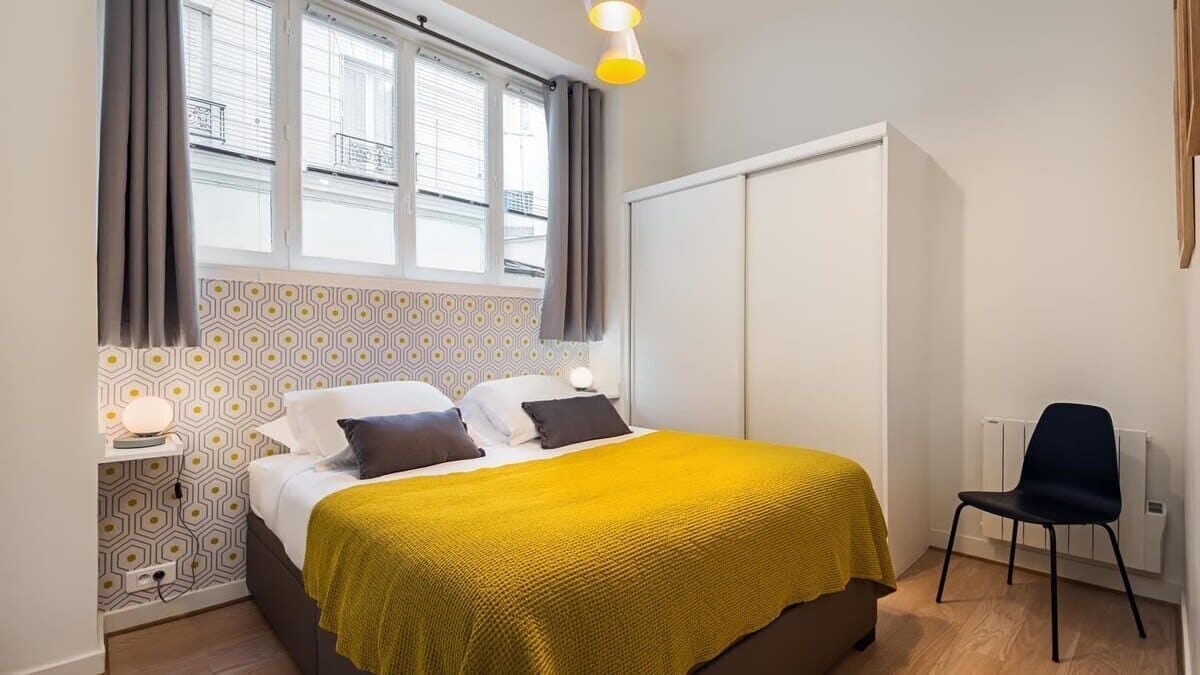 A bedroom with a bed, bedside table, lamp, chair, and a window with curtains.
