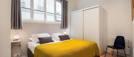 A bedroom with a bed, bedside table, lamp, chair, and a window with curtains.