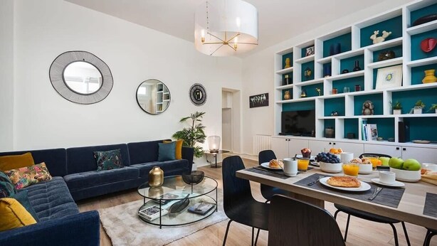 Una sala de estar moderna con un sofá azul, una mesa de centro de vidrio y un comedor con una mesa y sillas blancas. Hay espejos decorativos en la pared y una estantería llena de libros y objetos decorativos.