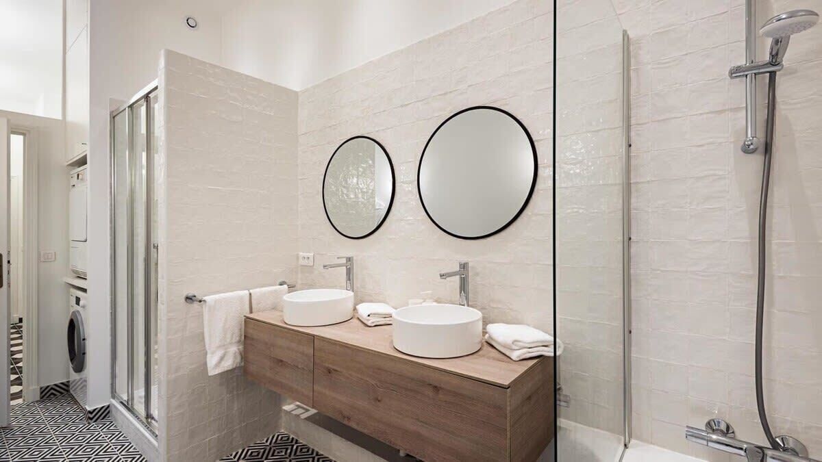 Un baño moderno con dos espejos redondos, un mueble de tocador de madera y una cabina de ducha con mampara de vidrio.