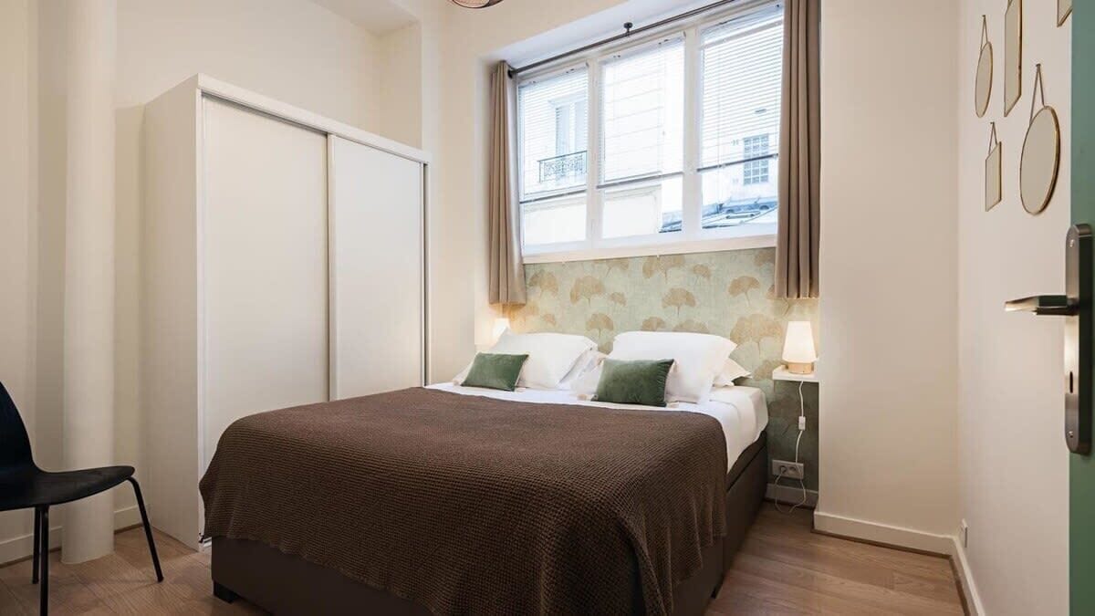 Ein Schlafzimmer mit Bett, Stuhl, Kleiderschrank, einem Fenster mit Vorhängen und Bildern an der Wand.