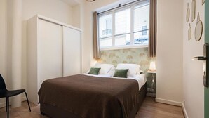 Un dormitorio con cama, una silla, un armario, una ventana con cortinas y una pared decorada con cuadros.