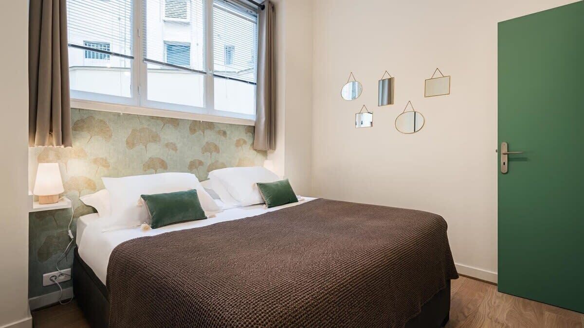 Une chambre d’hôtel avec un lit, une table de chevet, une lampe, une fenêtre avec des rideaux et une porte verte.