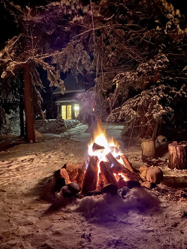 A snow-covered outdoor area with a campfire, trees, and a building in the background.