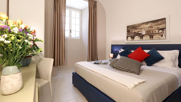 A bedroom with a bed, a bedside table with a vase of flowers, a window with curtains, and a painting on the wall.