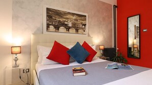 A hotel room with a bed, red and blue pillows, a framed picture on the wall, and a lamp on the nightstand.