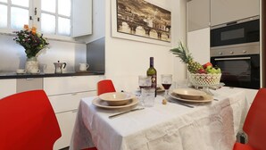 A dining area with a table set for two, a vase of flowers, a bottle of wine, and a fruit basket.