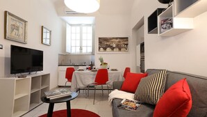 A modern living room with a flat-screen TV, a dining area with a table and chairs, and a grey sofa with red cushions.