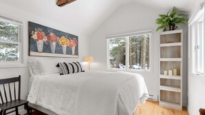 Une chambre avec un grand lit, une chaise en bois, une table de chevet avec une lampe, une fenêtre donnant sur des arbres et une étagère avec des bougies.
