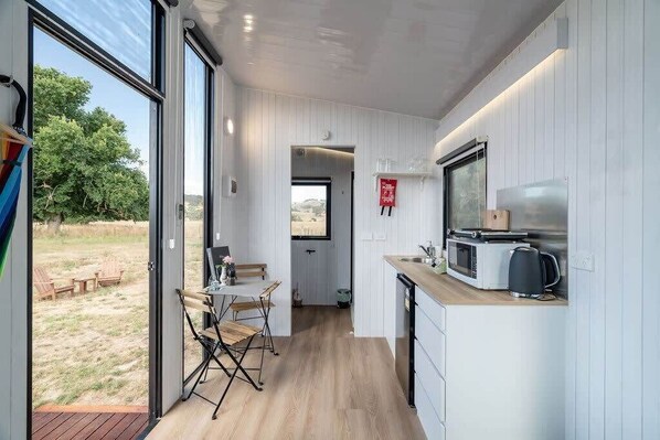 A compact kitchen with a wooden countertop, a microwave, and a kettle. A dining area with a table and chairs is visible through an open door. - JBJK Homestead by Tiny Away (Gobur)