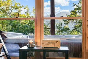 Quarto aconchegante com vista para árvores e montanhas através de grandes janelas. Uma mesa de cabeceira acomoda uma vela e uma placa de madeira com a palavra 'unplug'.