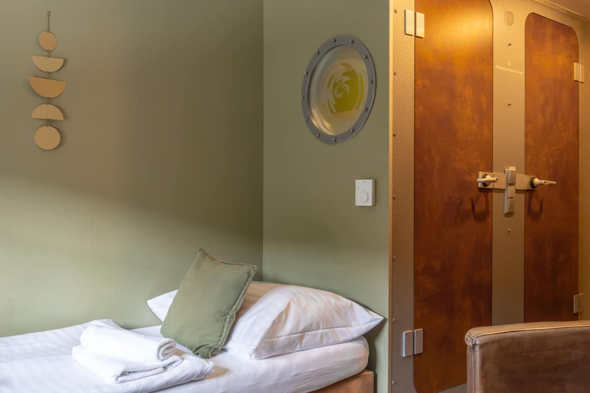 Una habitación con una cama, una silla, una mesa de madera, una lámpara de pared y una puerta con espejo.