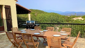 Een terras buiten met een houten tafel en stoelen, een barbecue en een prachtig uitzicht op heuvels en bergen.