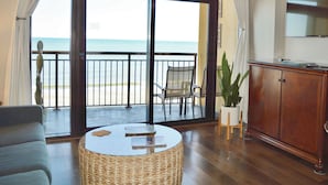 Ein Zimmer mit einer großen Schiebeglasdoor, die auf einen Balkon mit Meerblick führt. Es gibt einen Deckenventilator, einen runden Glastisch mit Büchern, einen Korbstuhl, einen Holzschrank und einen Blumentopf.