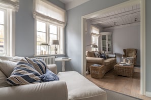 Uma sala de estar aconchegante com sofá, poltronas e uma pequena mesa. Há uma janela com cortinas, um radiador e um abajur. Uma cadeira e um cesto são visíveis ao fundo.
