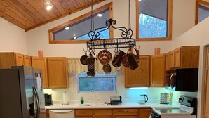 A kitchen with wooden cabinets, a stainless steel refrigerator, a dining area with a white table and wooden chairs, and a hanging pot rack above the island. - Riverfront Cabin w/ Porch & Firepit - Peaceful Retreat Near Murphy, NC. (Murphy)