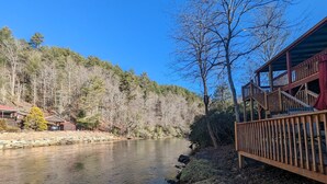 A cabin with a wooden deck overlooking a calm river, surrounded by dense forest. - Riverfront Cabin w/ Porch & Firepit - Peaceful Retreat Near Murphy, NC. (Murphy)