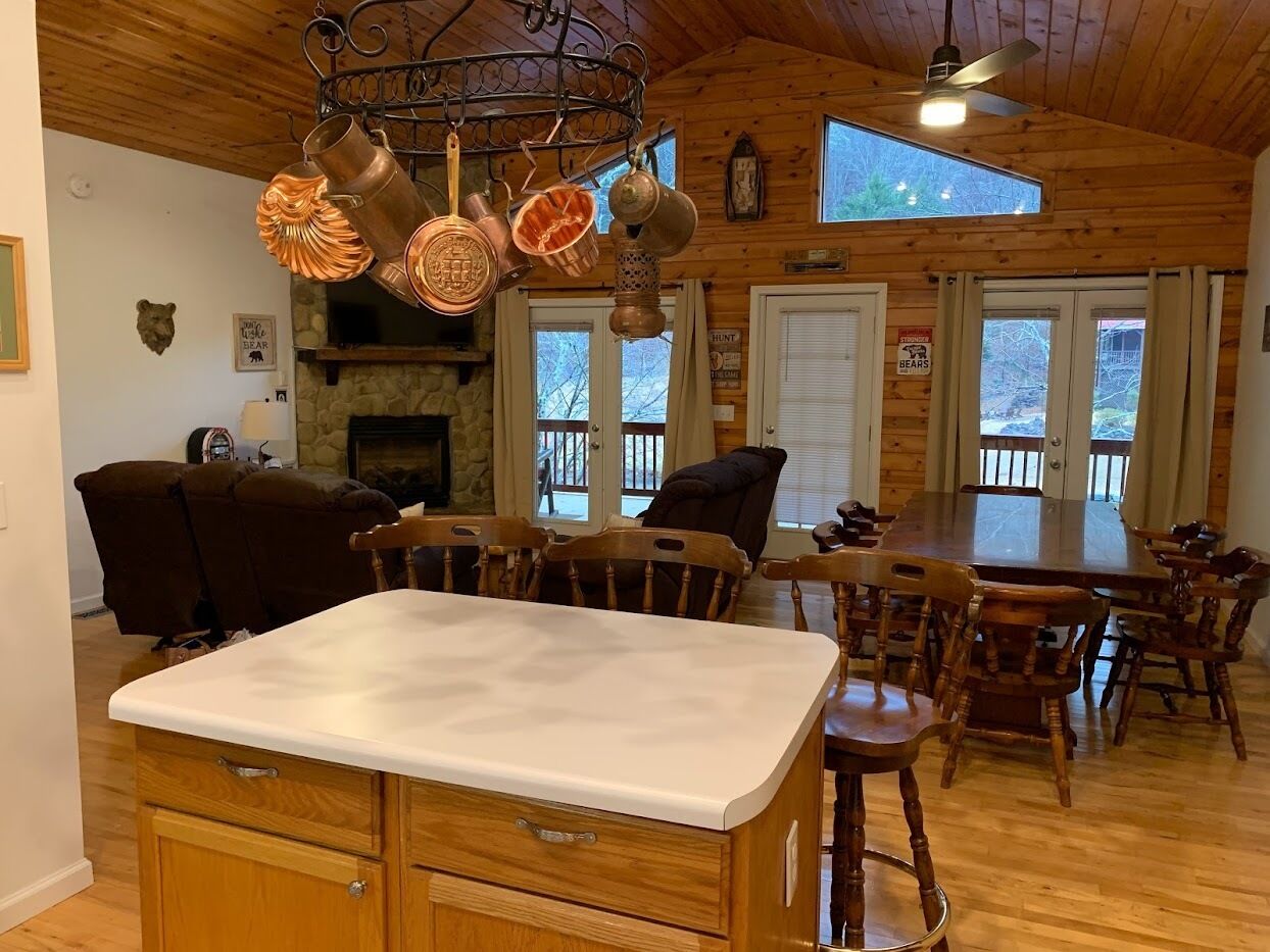 Un intérieur en bois avec un îlot de cuisine, une table à manger et des chaises. On y trouve des casseroles en cuivre suspendues au plafond, une cheminée en pierre et une grande fenêtre.