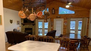 A wooden interior with a kitchen island, dining table, and chairs. There are copper pots hanging from the ceiling, a stone fireplace, and a large window. - Riverfront Cabin w/ Porch & Firepit - Peaceful Retreat Near Murphy, NC. (Murphy)