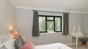 Un dormitorio con cama, almohadas, un escritorio con espejo, una lámpara y una ventana con cortinas.