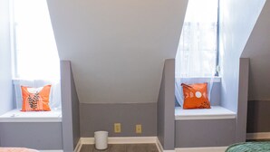 Une pièce avec un plafond en pente, deux fenêtres avec des rideaux, un seau blanc et deux lits recouverts de couvertures orange et vertes.