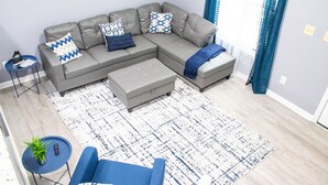 Un salon moderne avec un canapé d’angle gris, une petite table ronde, un fauteuil bleu et un tapis à motifs.