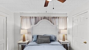 Ein Schlafzimmer mit einem weißen Bett, zwei Nachttischen mit Lampen, einem Deckenventilator und einem Fenster mit Vorhängen.