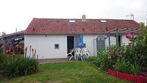 A single-story house with a red-tiled roof, a small porch with white plastic chairs, and a garden with various plants and flowers.