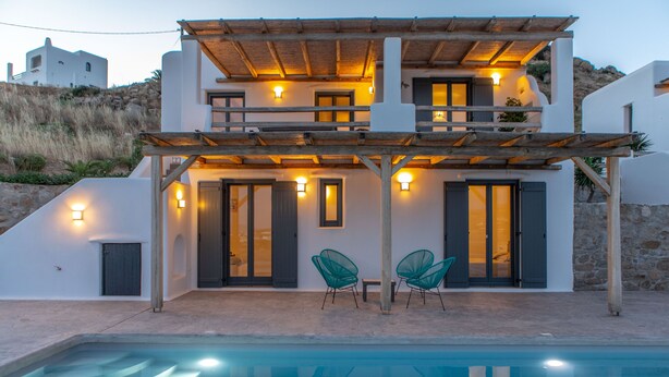 Ein modernes, zweistöckiges Haus mit Swimmingpool, Holz-Pergola und zwei grünen Stühlen.