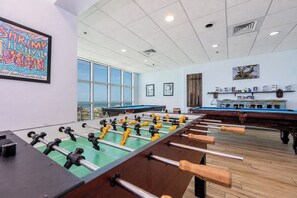 A room with two foosball tables, a pool table, and a large window providing a view of the beach.