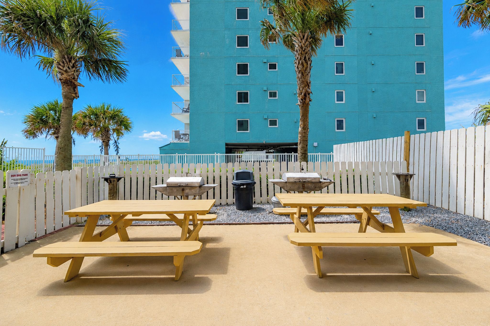 A beachfront patio with picnic tables, a trash can, and a white picket fence.