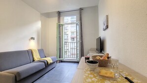 Ein gemütlicher Wohnbereich mit grauem Sofa, einem kleinen Esstisch mit gelbem Blumenläufer, einem Brotkorb, einem Glas Wasser und einem Fernseher an der Wand.