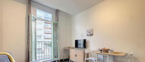 Quarto pequeno com porta de vidro deslizante para uma varanda, televisão em um armário, mesa com cadeiras e um pequeno quadro na parede.