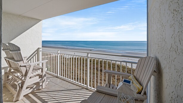 A balcony with a view of the beach, wooden chairs, and a small table with a cushion.