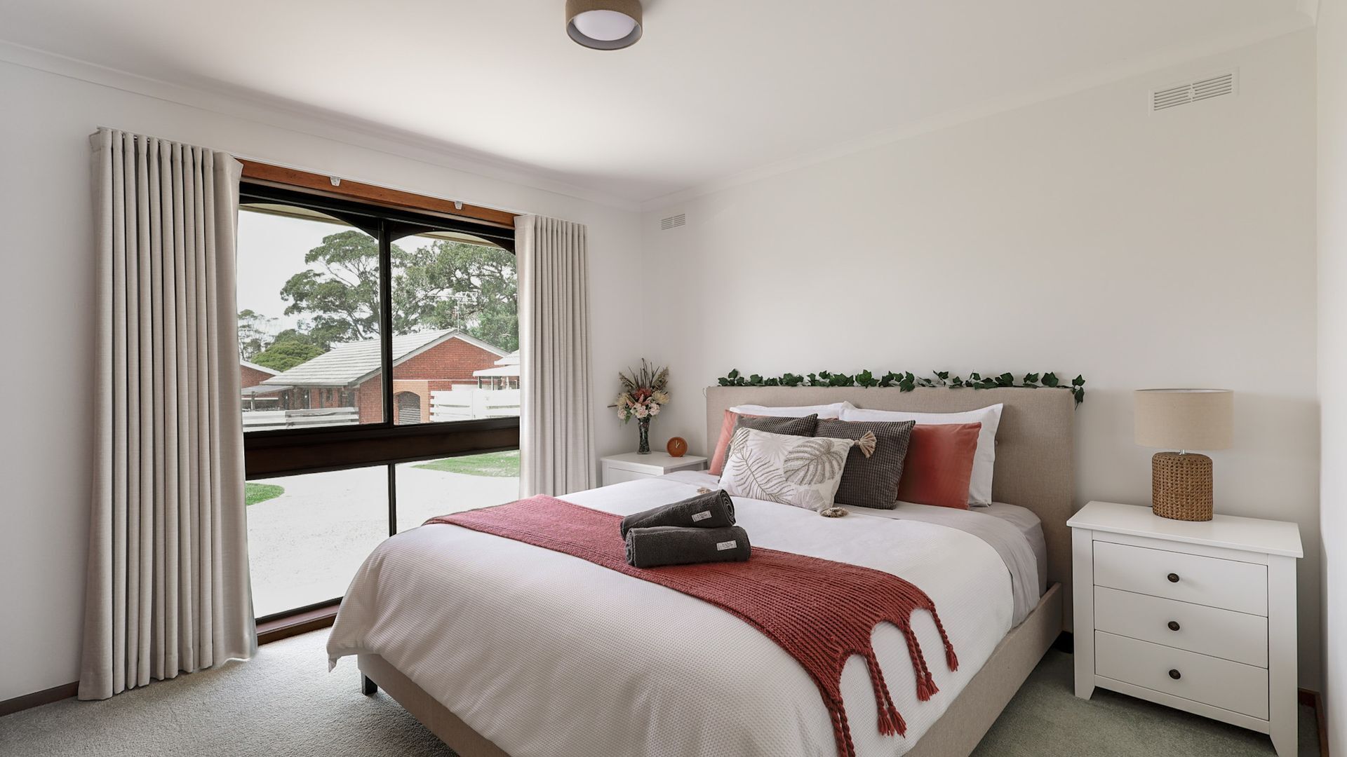 Een slaapkamer met een groot bed, witte nachtkastjes, een lamp en uitzicht op gebouwen.