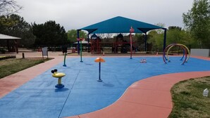 Ein Spielplatz mit einer blauen und roten Gummioberfläche, einer überdachten Spielanlage und einem eingezäunten Bereich.