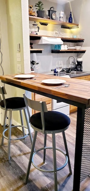A dining area with a wooden table, two black upholstered chairs, a kitchen in the background, and a white wall. - Cabin 14: Cute Rustic A-Frame Cozy Getaway w/ Kitchen (Glenwood Springs)