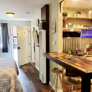 Ett kompakt bostadsutrymme med ett litet kök, en säng med mönstrat sänglinne och en matplats med pallar.