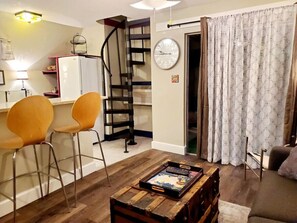A living room with a wooden coffee table, two yellow chairs, a spiral staircase, a clock, and a patterned curtain. - Cabin 17: Cozy 2-Story Retreat w/ Spiral Staircase • Kitchen • Sleeps 4 (Glenwood Springs)