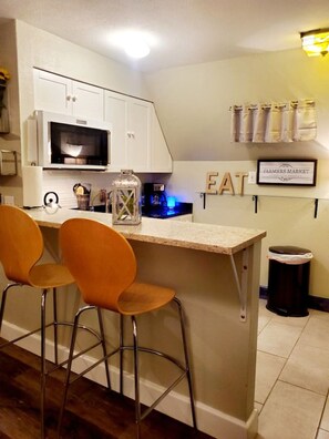 Una cocina con barra, armarios blancos, microondas, cafetera y un bote de basura.