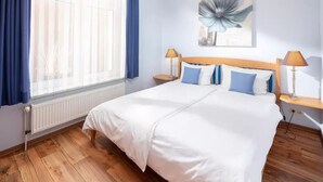 A bedroom with a wooden floor, a bed with white and blue bedding, two bedside tables with lamps, and a large flower painting on the wall. - Starfish, city center, central location, wifi, very quiet (Norderney)
